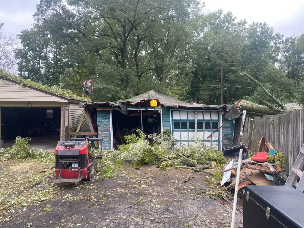 A damaged blue garage in Indianapolis, IN with a fallen tree crushing the roof, debris scattered around, and a worker on top making repairs. McMahon Rolloffs provides storm damage junk removal and waste hauling services in Indianapolis for fast cleanup after disasters.